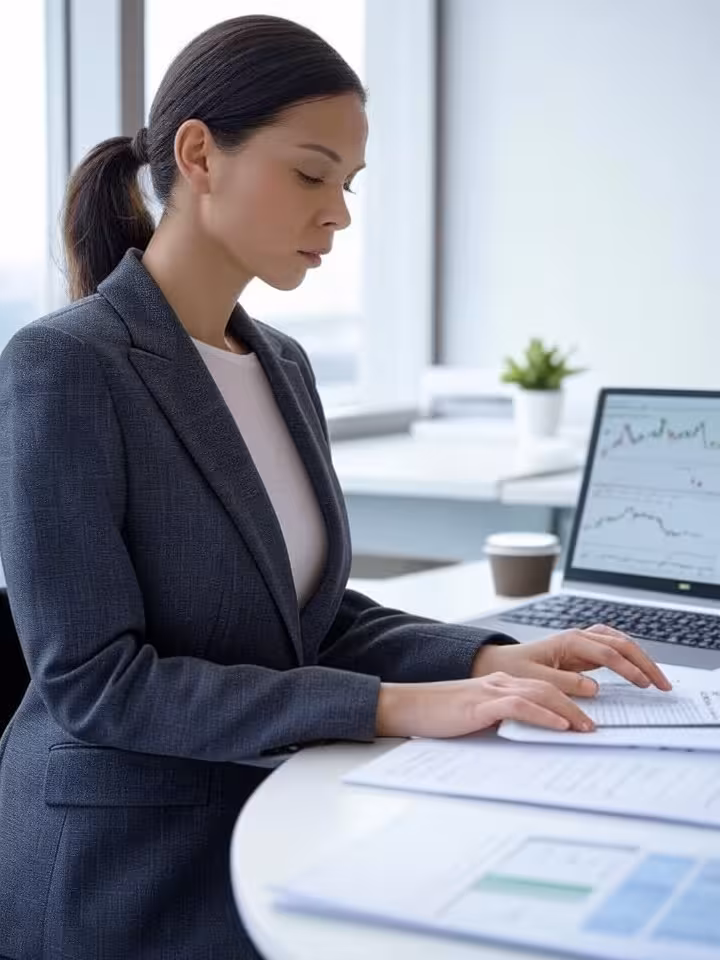 Professional woman reviewing documents on laptop at modern workspace, financial charts visible on sc